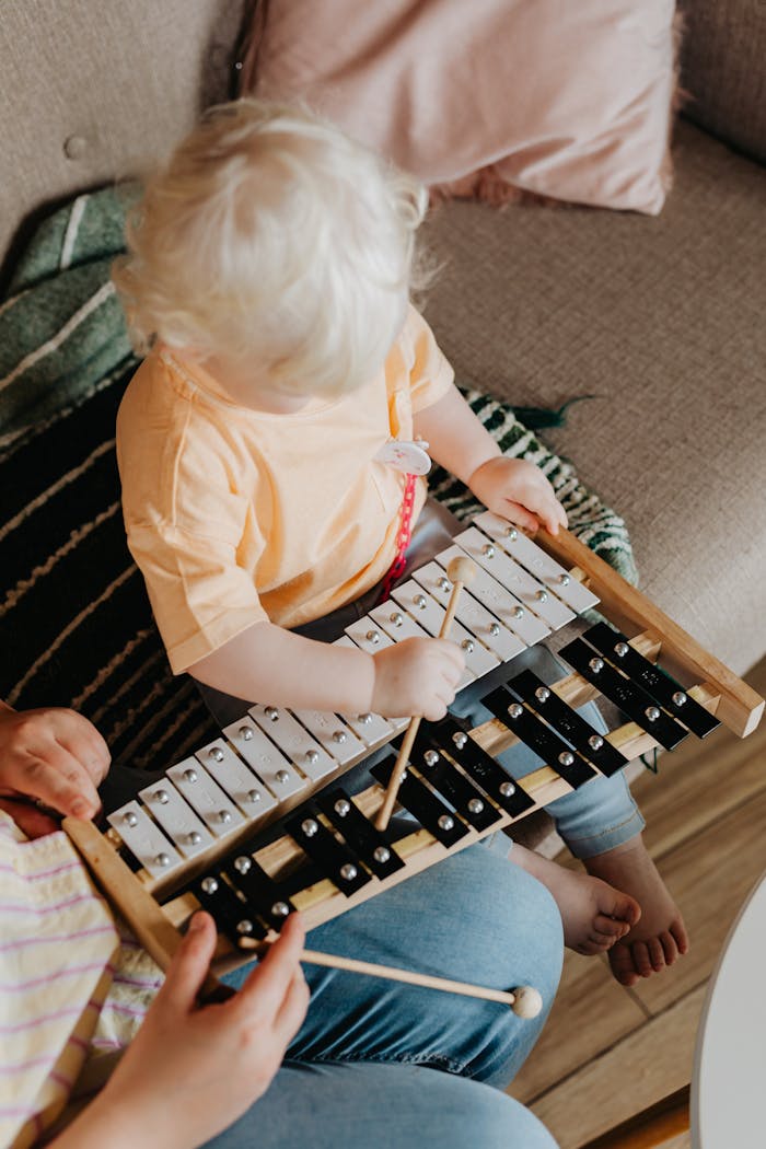A young child sitting on a sofa plays a xylophone with adult supervision.