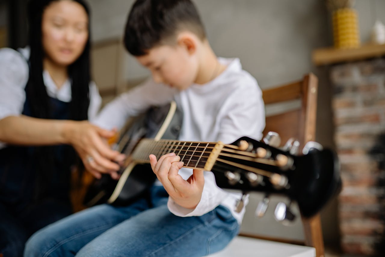 Young boy learning guitar indoors with guidance, focus on fretboard.